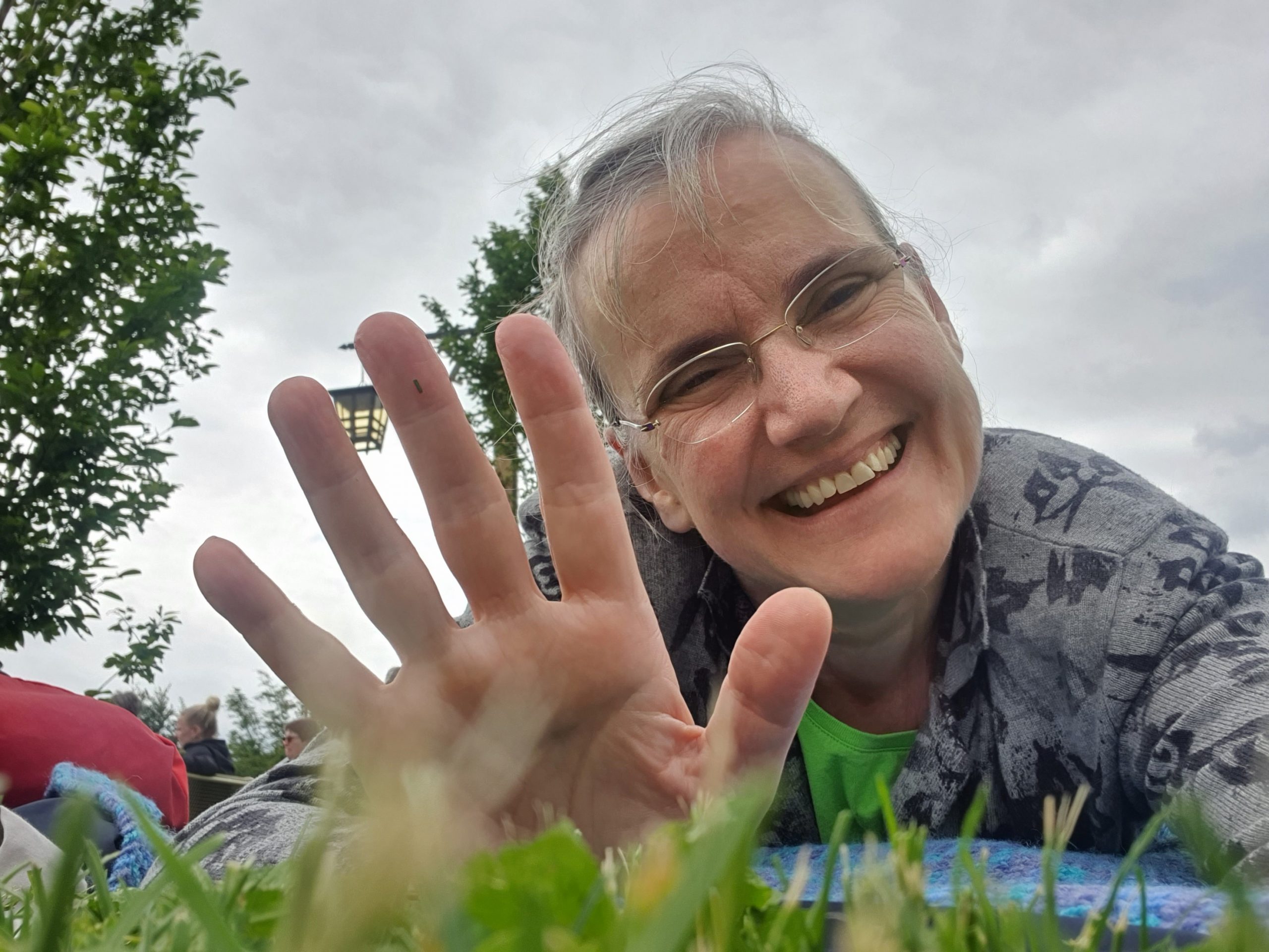 Ich liege im Gras auf dem Bauch und lächle glücklich mit winkender Geste.